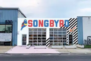 Storefront of Songbyrd, featuring bold, colorful signage with a bird illustration, large windows, and a black-and-white striped mural detail on a city street.